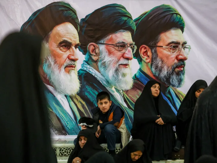 Oameni ascultând discursuri în fața unui mural cu lideri iranieni.