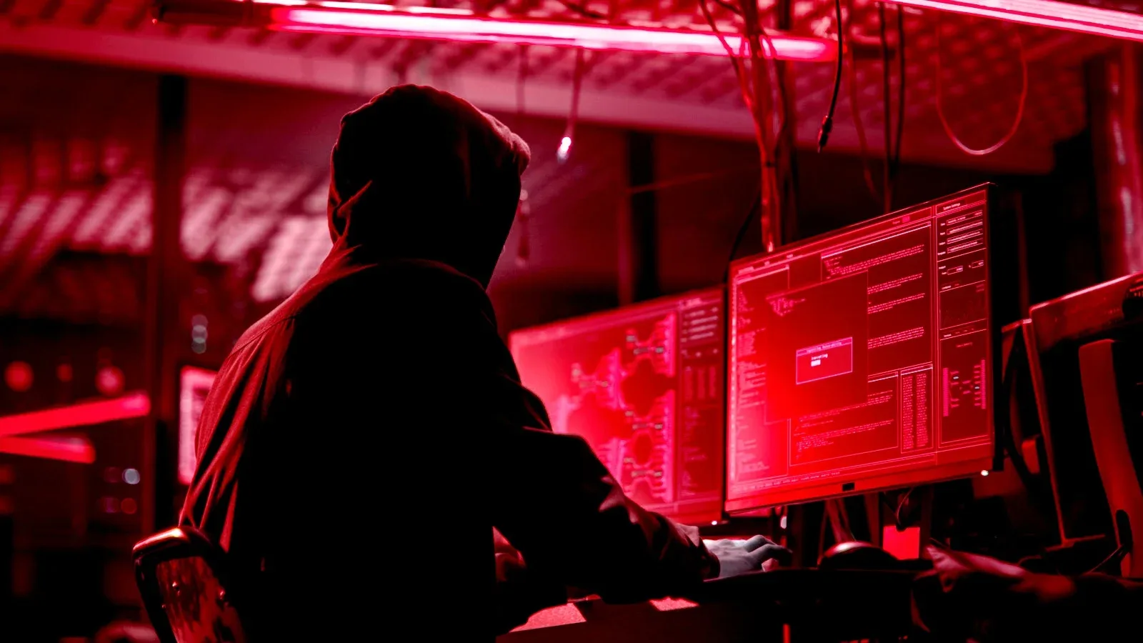 Un hacker lucrează la computere în lumină roșie, evidențiind riscurile cibernetice.