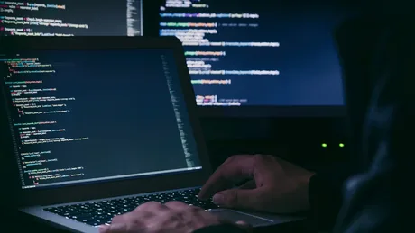 Un atacator lucrează la un laptop, coduri pe ecranul din fundal.