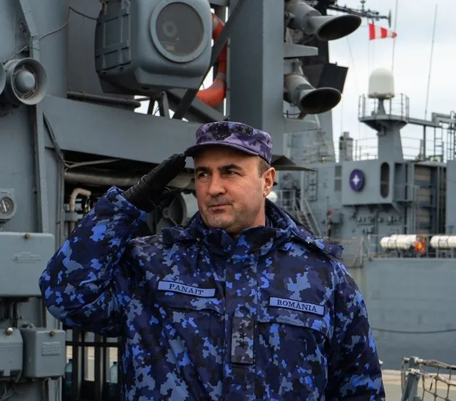 Oficial militar român în uniformă, salutând pe fundalul unei nave de război.