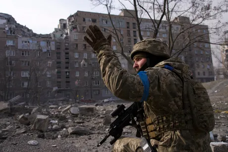 Soldat ucrainean în fața unei clădiri distruse, semnalizând cu mâna.