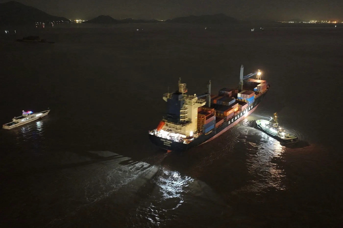 O navă de containere navighează pe ape întunecate, cu lumini strălucitoare pe punte.