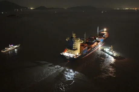 O navă de containere navighează pe ape întunecate, cu lumini strălucitoare pe punte.