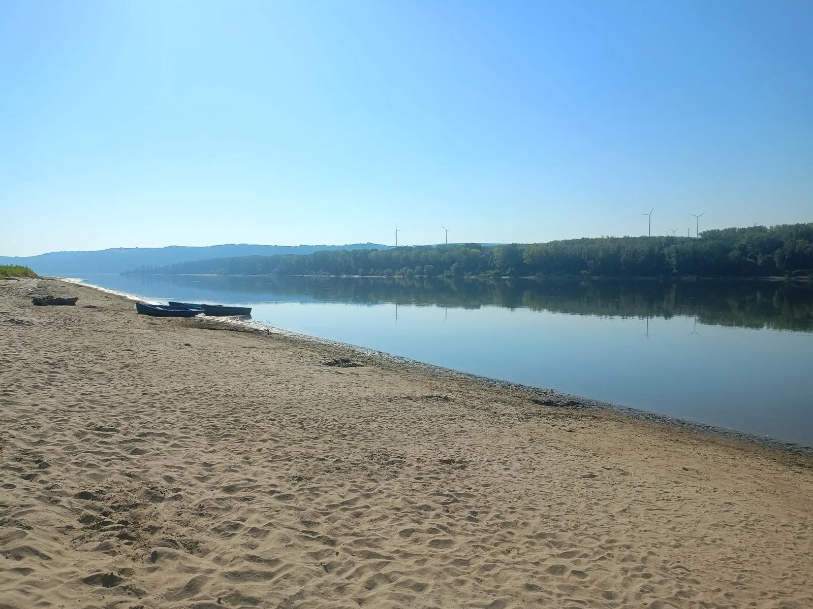 Plajă liniștită pe malul Dunării, cu apă calmă și vegetație în fundal.