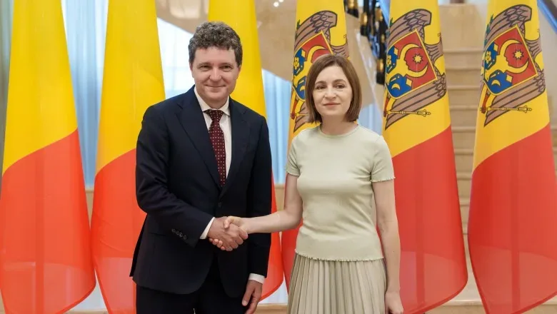 Maia Sandu și un oficial român strâng mâinile în fața drapelului.