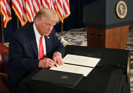 Donald Trump semnează documente în fața steagurilor americane.