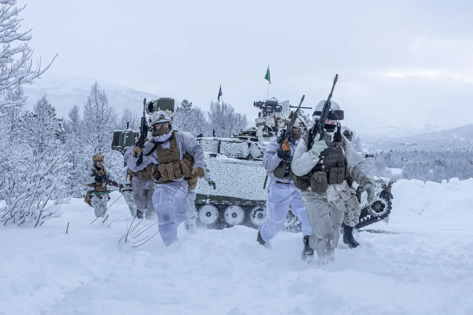 Soldați în echipament de iarnă desfășurându-se în zăpadă, cu vehicul militar în fundal.
