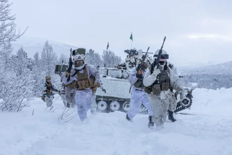Soldați în echipament de iarnă desfășurându-se în zăpadă, cu vehicul militar în fundal.