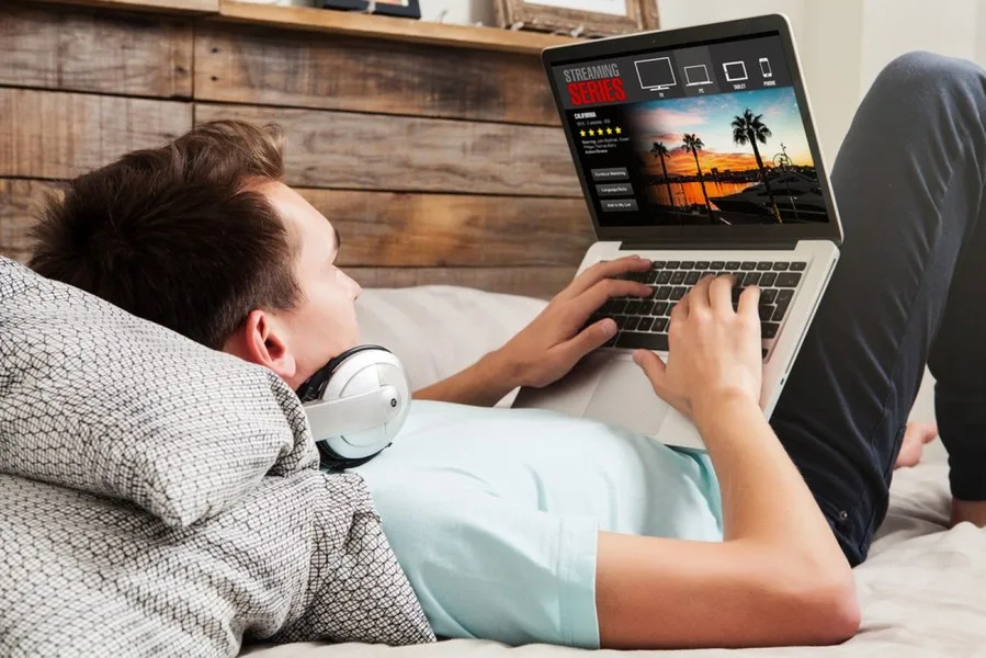 Un tânăr stă întins pe pat, folosind un laptop pentru a viziona un serviciu de streaming.