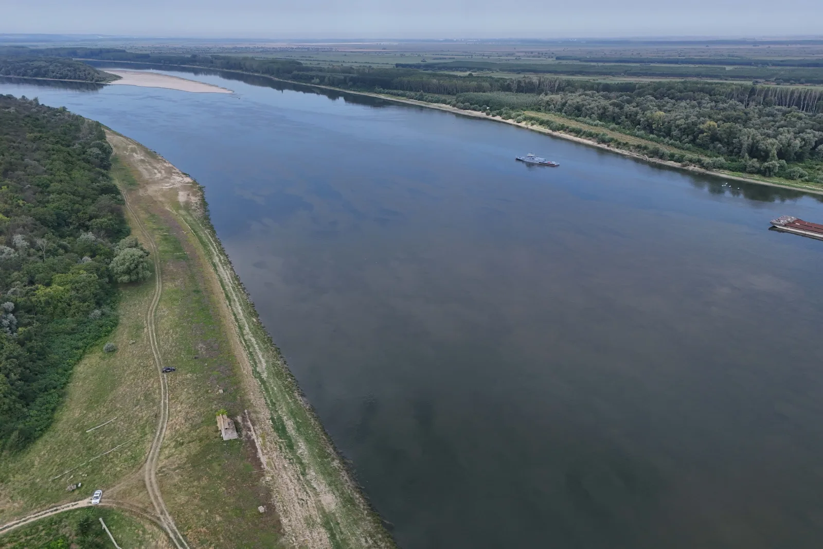 Dunărea cu maluri verzi și apă liniștită, barje în depărtare.