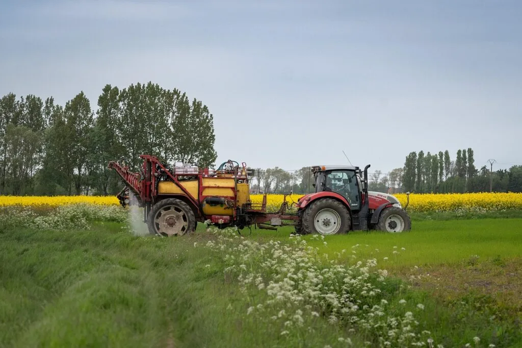 Tractor lucrând pe câmpuri verzi, cu flori galbene în fundal.