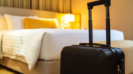 O valiză neagră lângă un pat de hotel confortabil, pregătită pentru oaspeți.