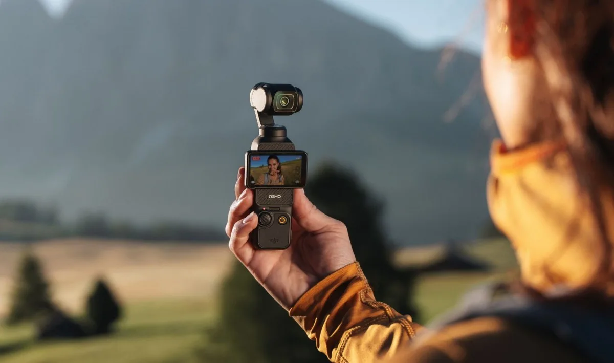 O persoană ținând o cameră compactă de vlogging în natură, cu peisaj montan în fundal.