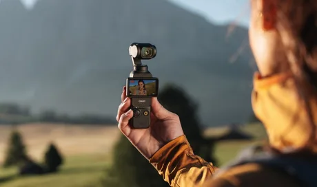 O persoană ținând o cameră compactă de vlogging în natură, cu peisaj montan în fundal.