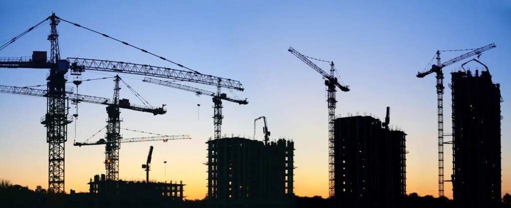 Silhouette de clădiri în construcție la apus, cu macarale vizibile.