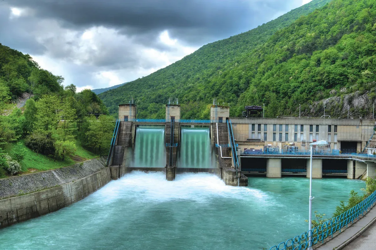 Baraj hidroelectric cu apă curgând, înconjurat de natură verde.