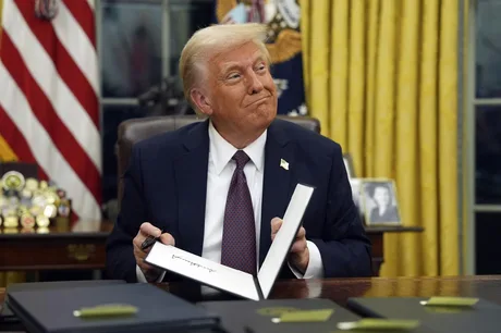 Donald Trump semnează documente în biroul oval, cu un zâmbet pe față.