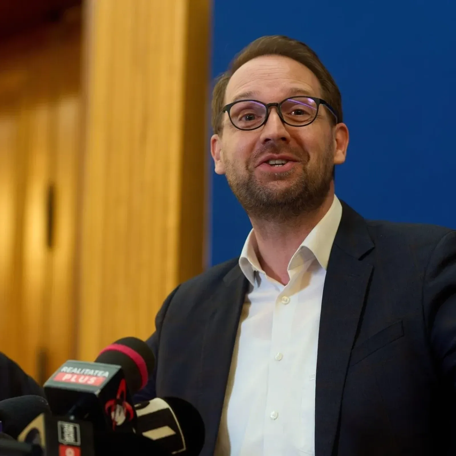Dominic Fritz zâmbește în fața presei, cu microfoane în jur.