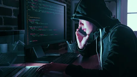 Un hacker lucrează la un computer, folosind un telefon mobil pentru comunicare.