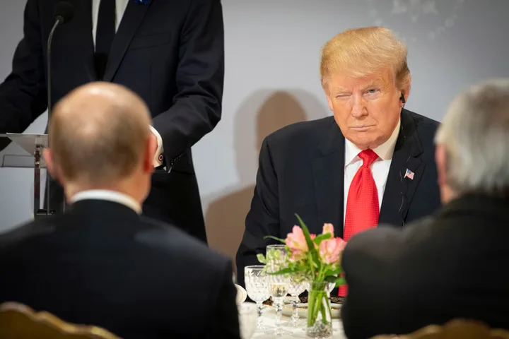 Donald Trump ascultând discursuri într-o întâlnire oficială, cu un zâmbet enigmatic.