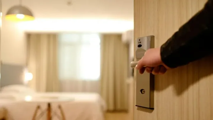 Ușa unei camere de hotel deschisă, cu un pat și perdele în fundal.