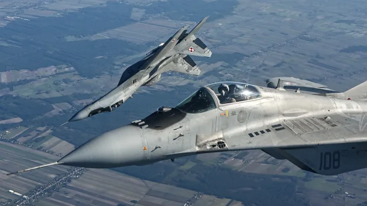 Două avioane MiG-29 zburând în tandem pe un fundal de câmpuri verzi.