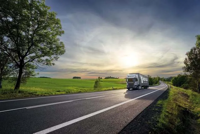 Camion pe drum, peisaj rural cu cer senin și soare apunând.