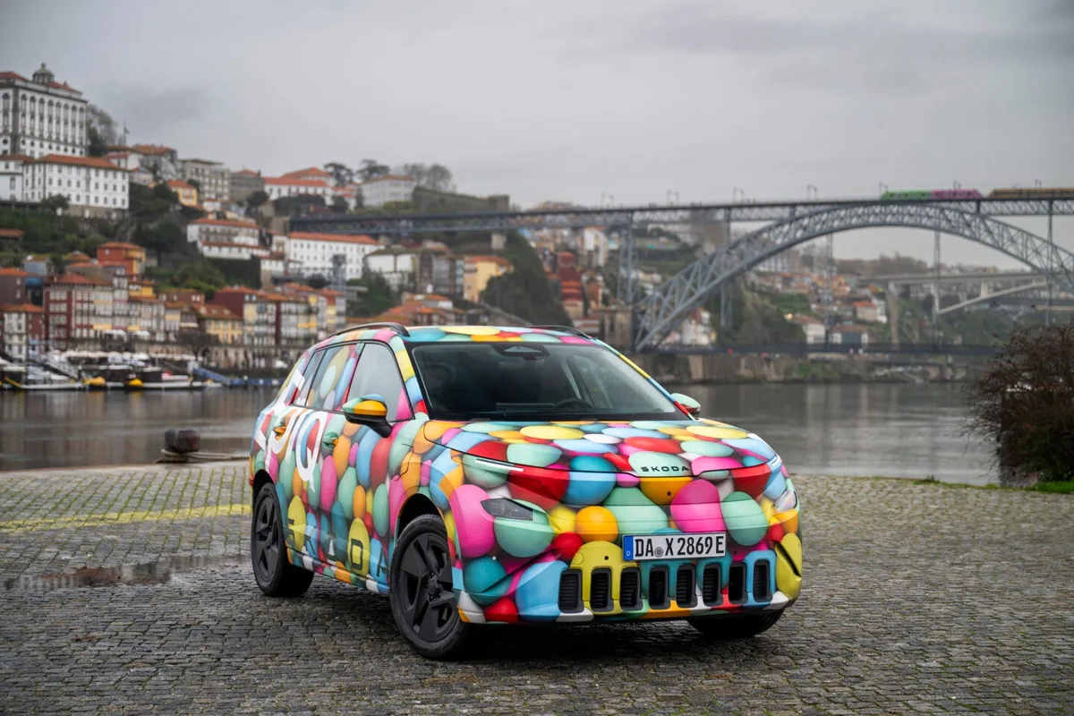 Skoda Epiq camuflat pe malul unui râu, cu un peisaj urban în fundal.