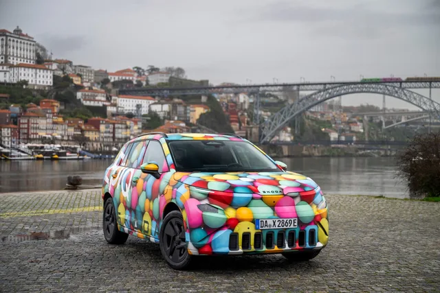 Skoda Epiq camuflat pe malul unui râu, cu un peisaj urban în fundal.