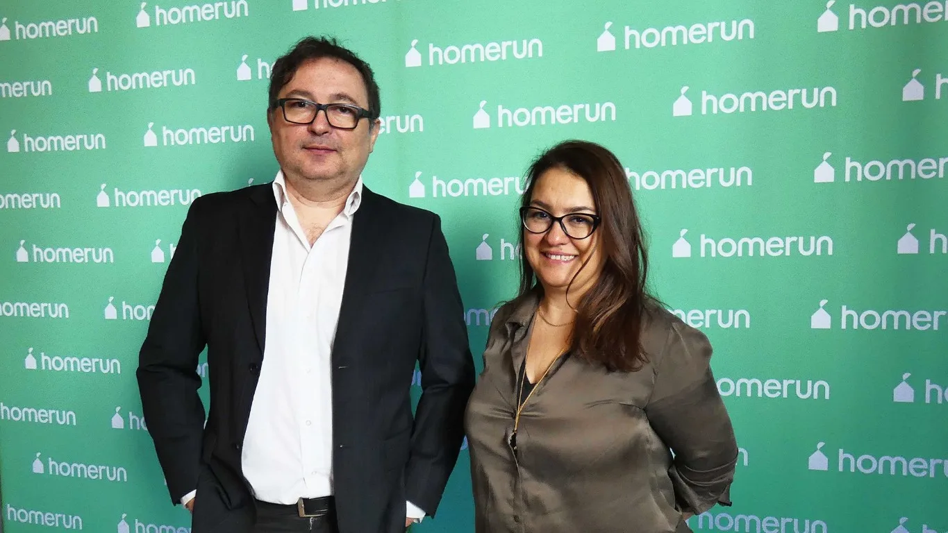 Două persoane zâmbind, în fața unui fundal verde cu logo-ul HomeRun.