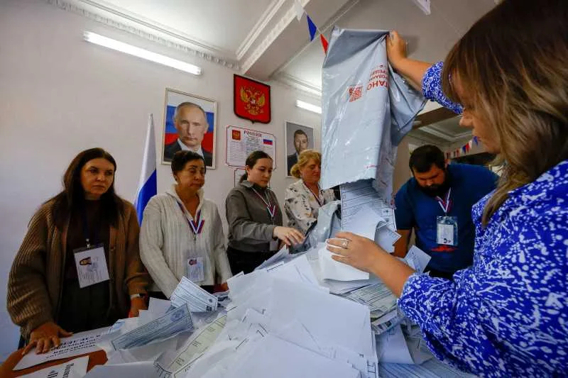 Oameni la un birou electoral în timpul alegerilor din Rusia, cu buletine de vot pe masă.