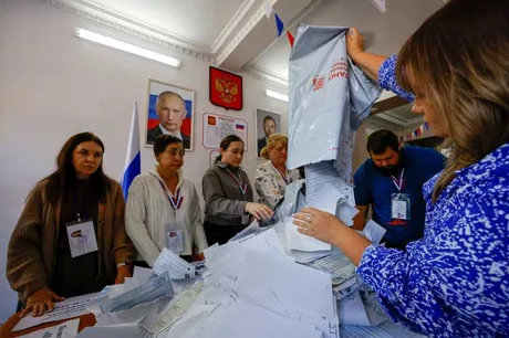 Oameni la un birou electoral în timpul alegerilor din Rusia, cu buletine de vot pe masă.
