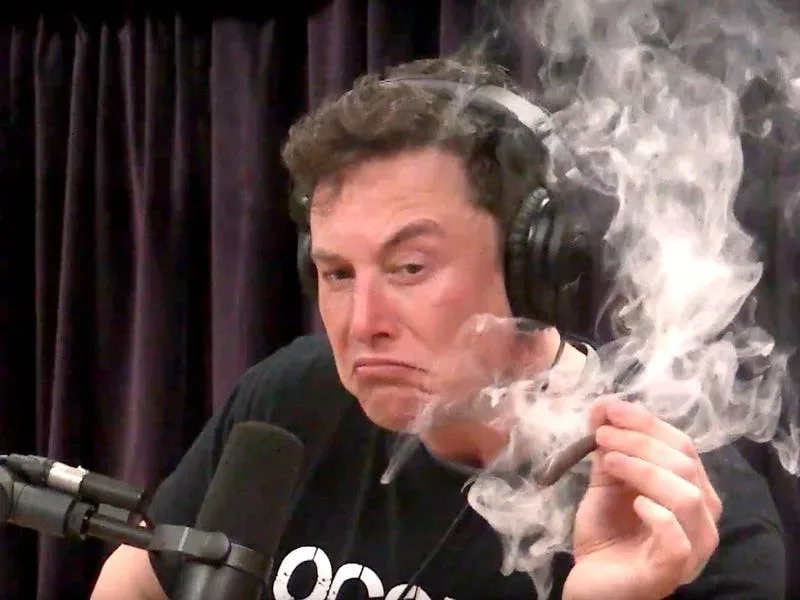 Elon Musk discutând în studio, cu un fum de țigară în jur.