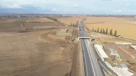 Peisaj al șantierului Autostrăzii A7, cu lucrări în desfășurare și teren agricol în fundal.