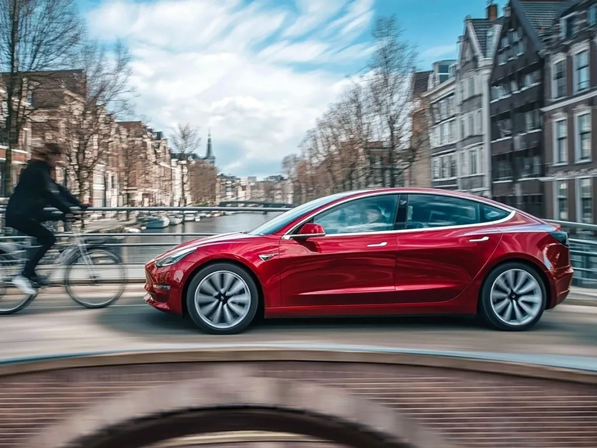 Un automobil Tesla roșu circulând pe un pod în Olanda, cu un ciclist în fundal.
