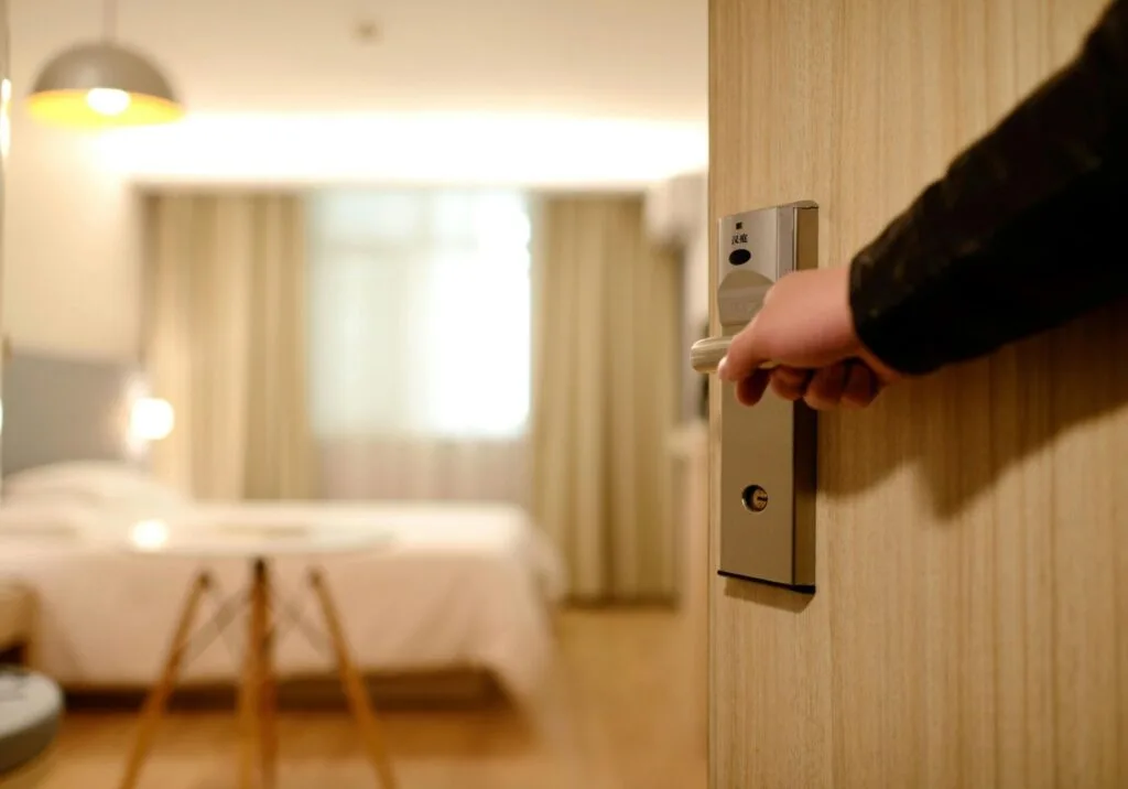 O mână deschide o ușă către o cameră de hotel modernă și luminoasă.
