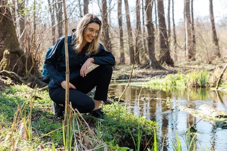 Diana Buzoianu zâmbește lângă un pârâu, înconjurată de natură.